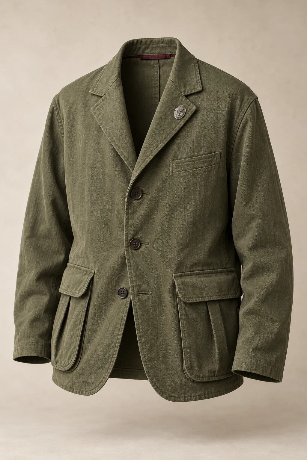 An olive herringbone field blazer with soft lapels, three buttons, bellows pockets and a small silver oval lapel pin.